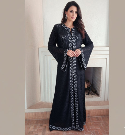 Woman wearing a black moroccan kaftan dress with white patterns in a room with a fireplace.