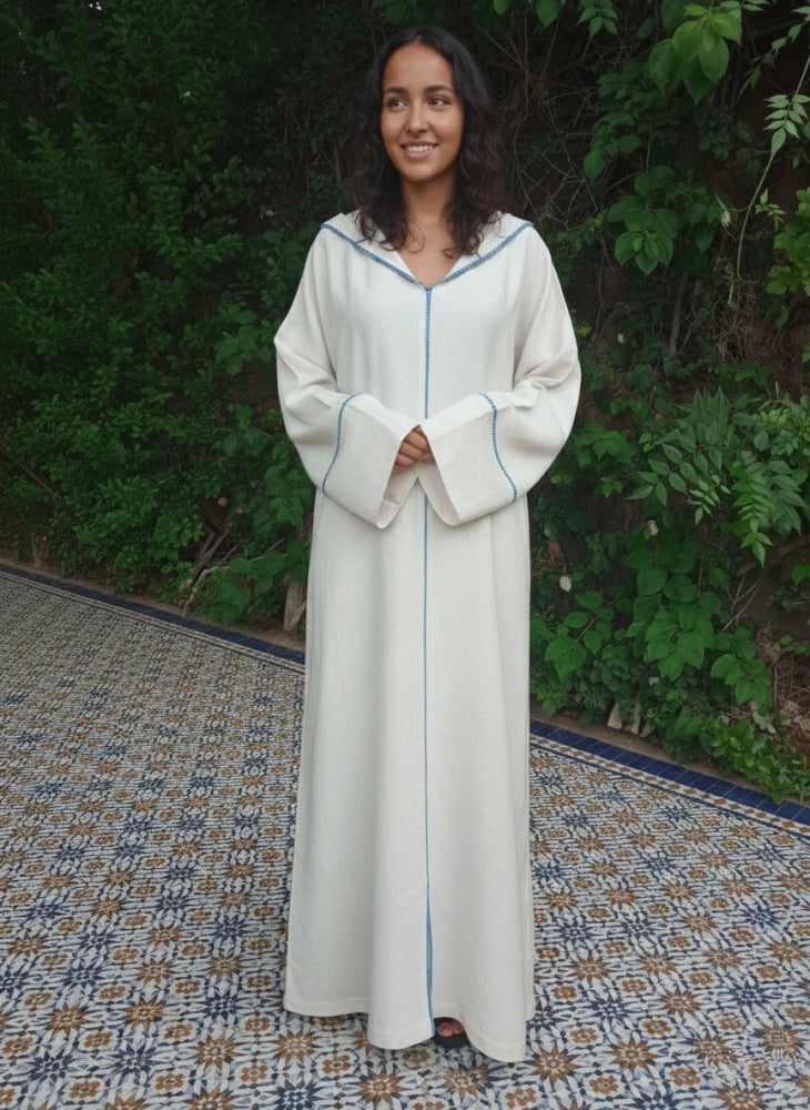 Woman wearing a long, light-colored Moroccan djellaba dress with a blue trim against a green outdoor background.