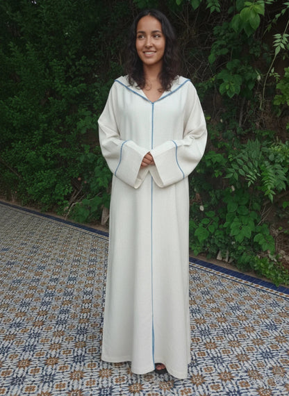 Woman wearing a long, light-colored Moroccan djellaba dress with a blue trim against a green outdoor background.