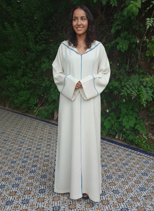 Woman wearing a long, light-colored Moroccan djellaba dress with a blue trim against a green outdoor background.