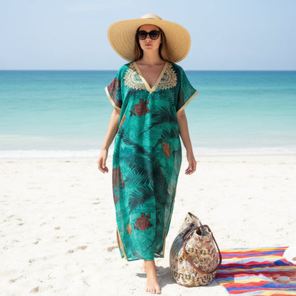 Woman in a green beach gandoura dress and straw hat standing on a sandy beach with ocean in the background.
